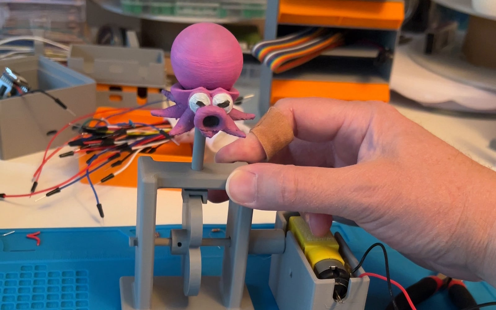 A hand turning the crank on a grey 3D-printed automata frame, with a painted magenta Octorok riding the shaft on top and a small yellow gearmotor sitting alongside it on the bench.
