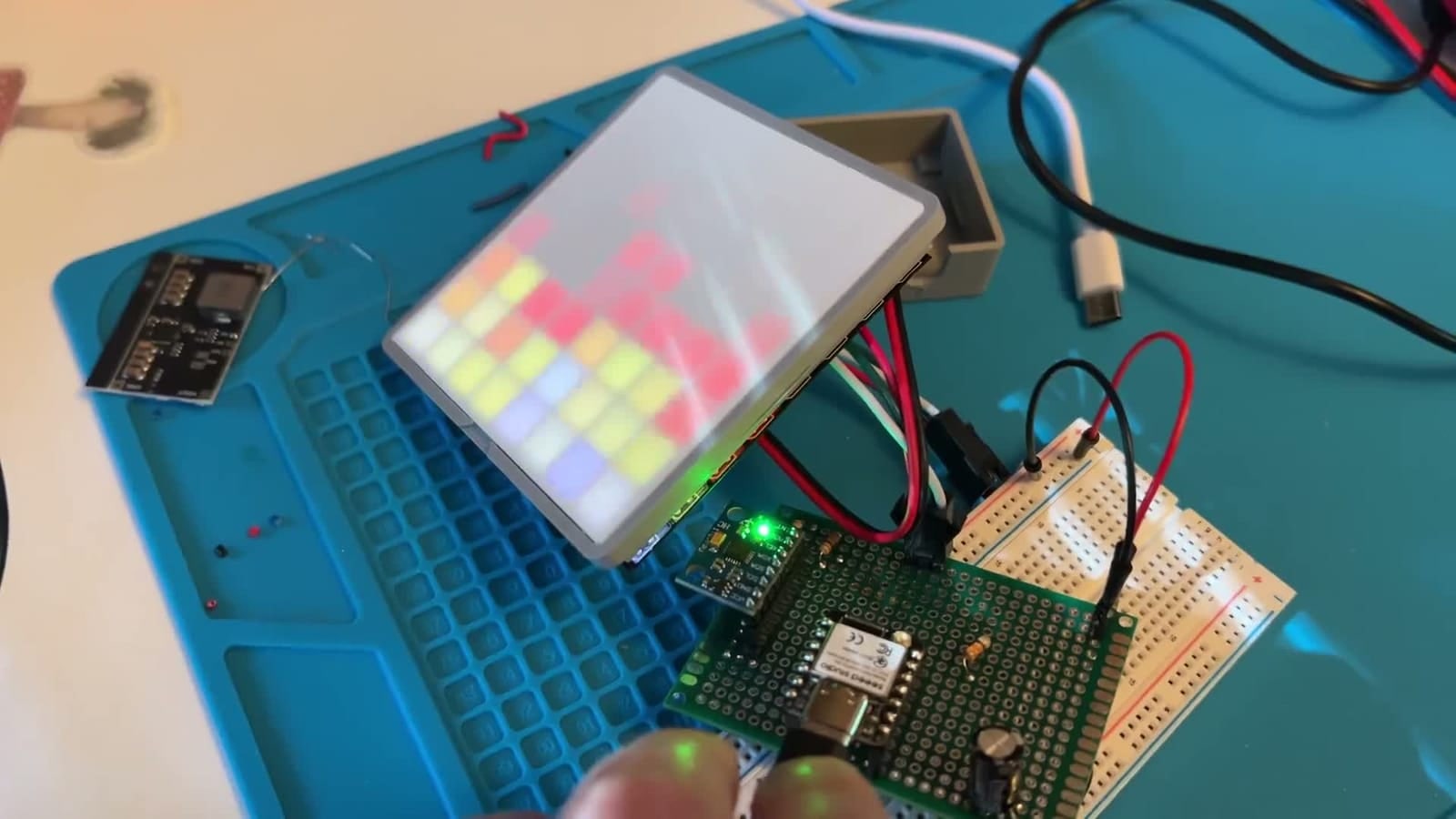 A small LED tile propped on a blue workbench mat behind a white diffuser, its grid glowing in soft reds, yellows and lavenders, wired to an ESP32 on protoboard and a breadboard.