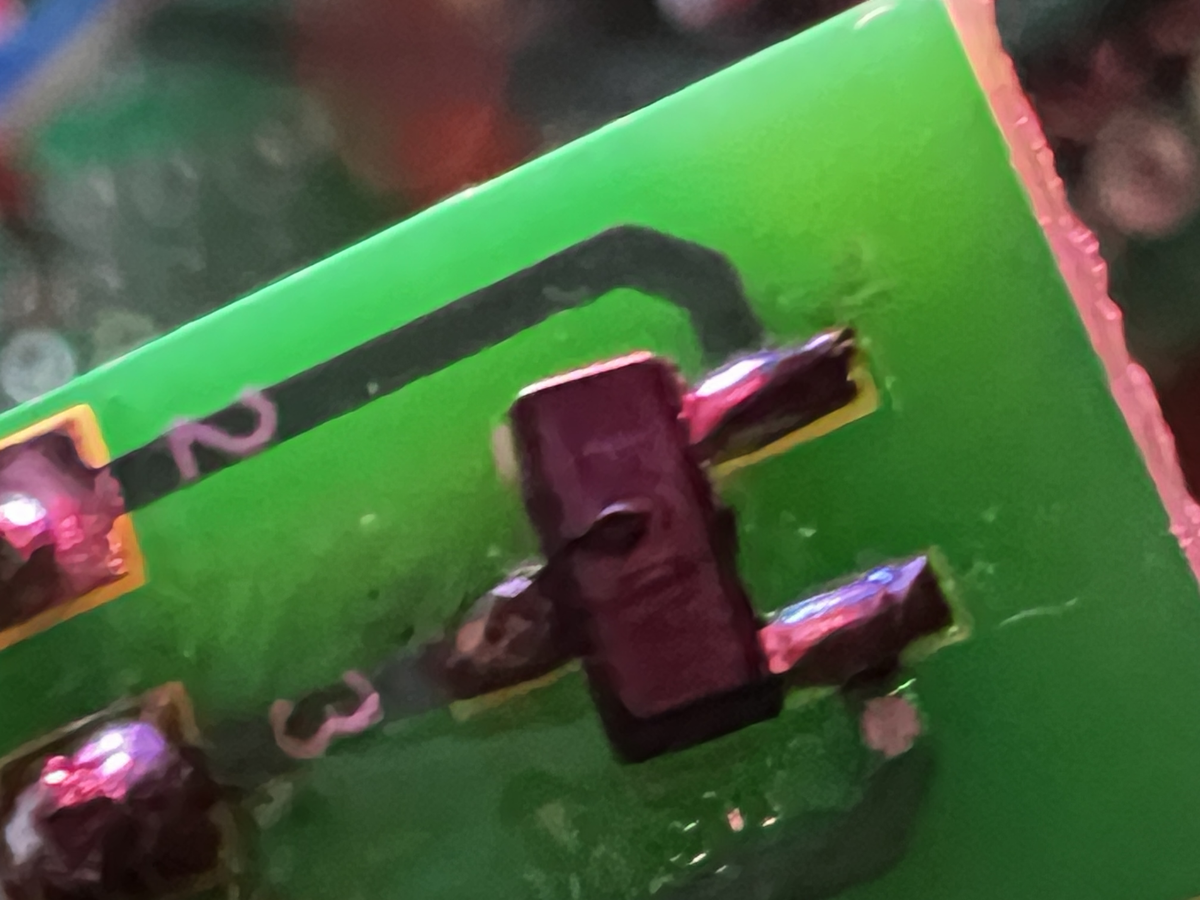 Extreme close-up of a tiny black three-pin surface-mount component soldered to a green breakout board, lit pink, with a small raised bubble on its top face.