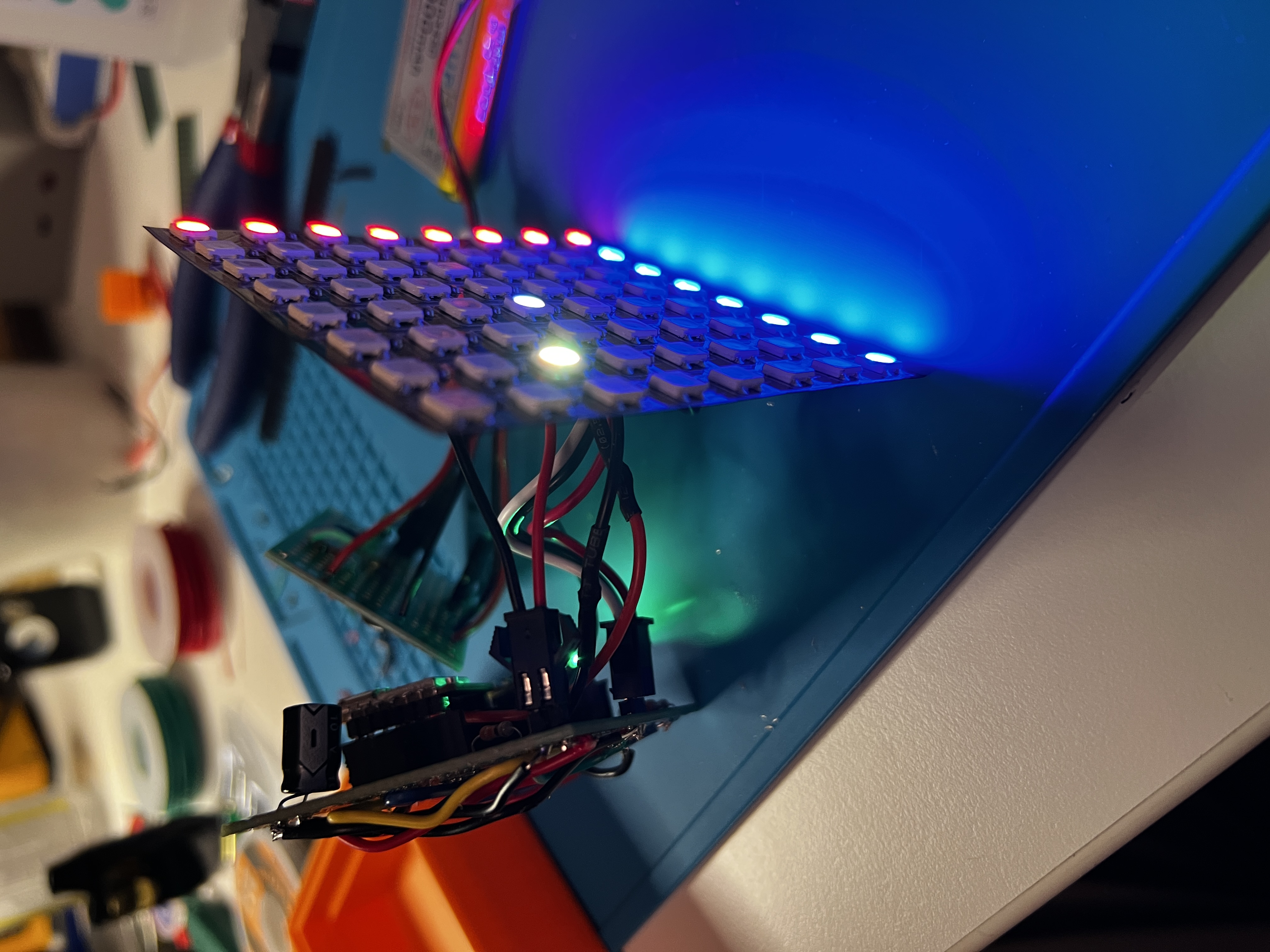 A flexible 8x8 LED panel propped up on a workbench, its top row glowing red and the rest violet, blue and cyan with one bright white pixel, throwing blue light across the surface behind it.