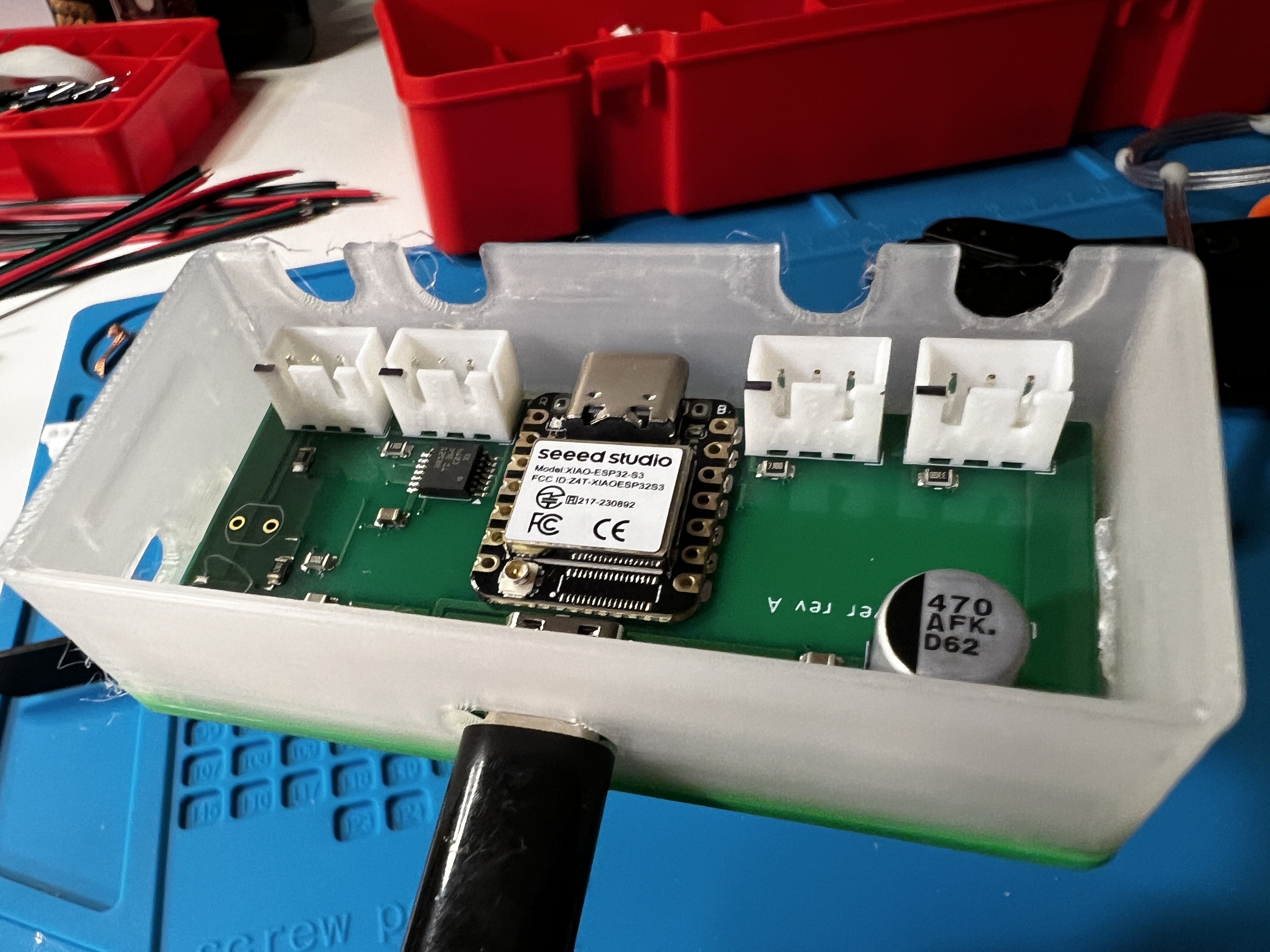 A green rev A circuit board sitting in a translucent white 3D-printed tray on a blue work mat, carrying a Seeed Studio XIAO module, four white connector sockets along one edge and a silver capacitor, with red parts bins behind it.